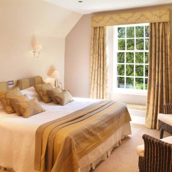 a picture of a cream bedroom, with gold and light brown accents in curtains, cushions and headboards, with a view of green trees out of the window