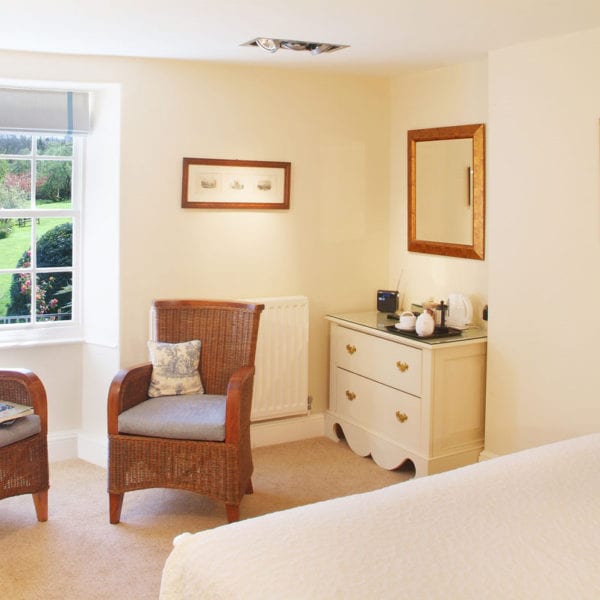 a picture of the view out of a cream bedroom. two brown wicker chairs with one stacked with magazines, the behind view a garden with green trees and patio furniture