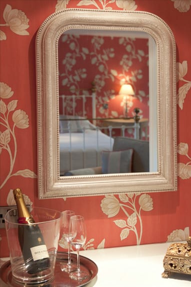 A picture of a mirror reflecting a bedroom view, red wallpaper with cream flowers and a bottle of champagne