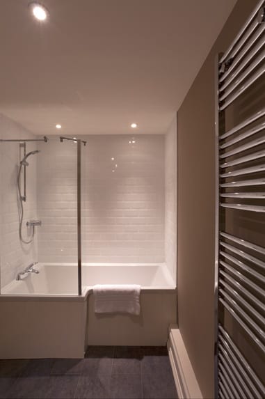 a picture of a white brick bathroom, with a wide set white square bath, a white towel and an overhead silver coloured shower