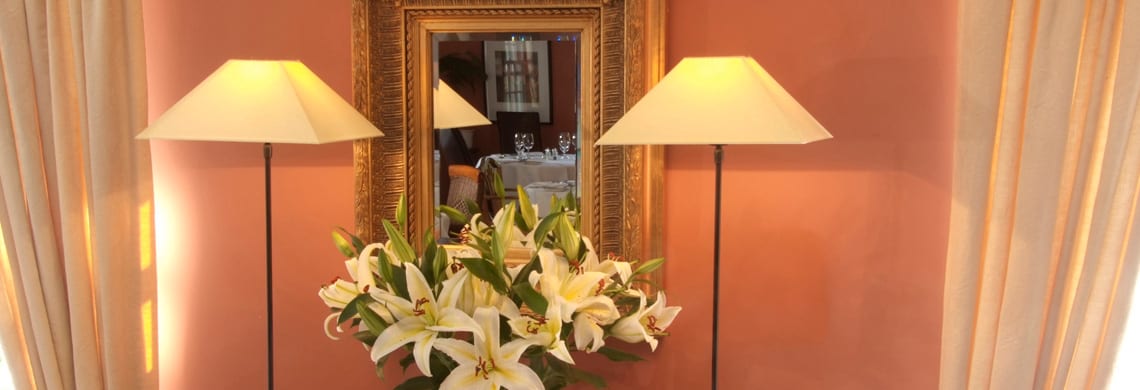 a picture of an orange dining room with cream curtains, two lamps, a wooden framed mirror and a vase of white lilies