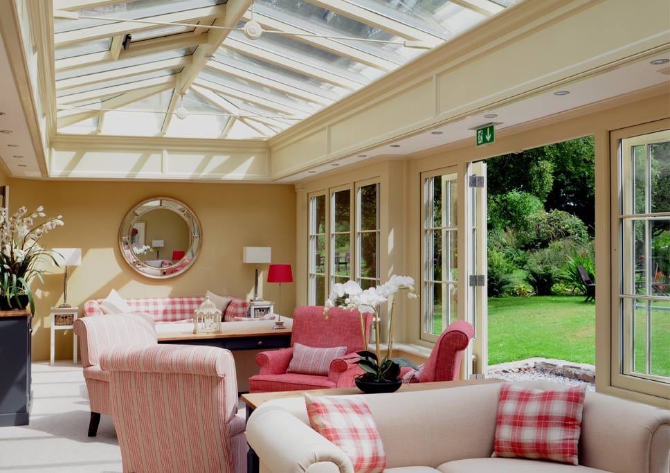 a picture of a cream dining and relaxing room with a glass roof, various sofas and patio doors that open up onto a green garden