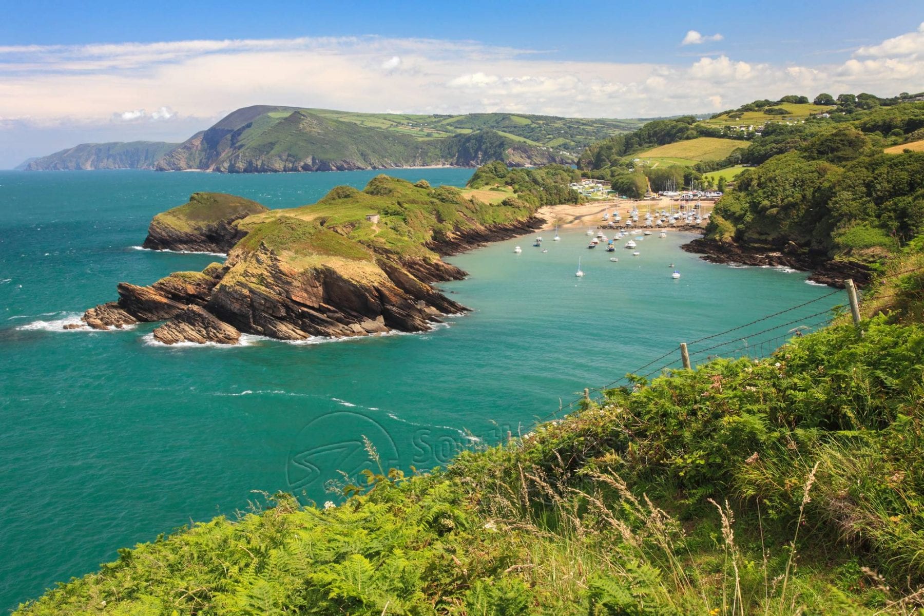 a picture of Watermouth coastal path with boats resting on the blue sea and green rocky cliffs surrounding the cove