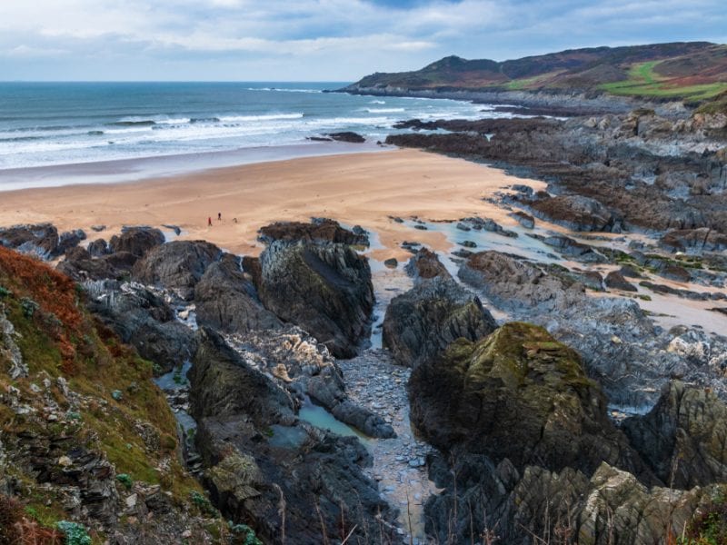 A picture of Barricane beach, colourful rocks surrounding a sandy beach with the sea in the background