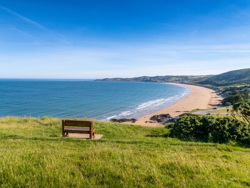 A picture of a landscape at Putsborough with a bench on the grass, over looking the coast and hills
