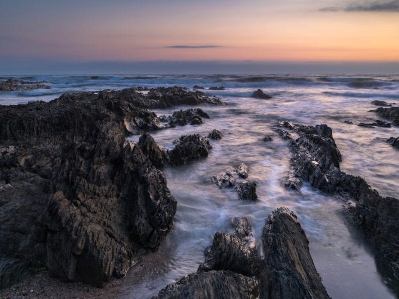 a picture of the flowing sea amongst slate grey rocks with a sunset in the background