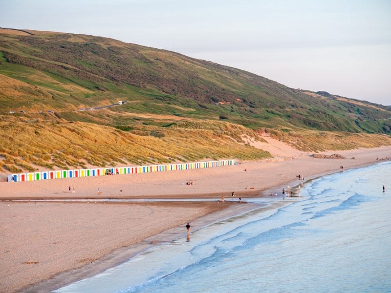 a picture of Woolacombe, landscape of green hills, colourful beach huts, sand and the light blue sea