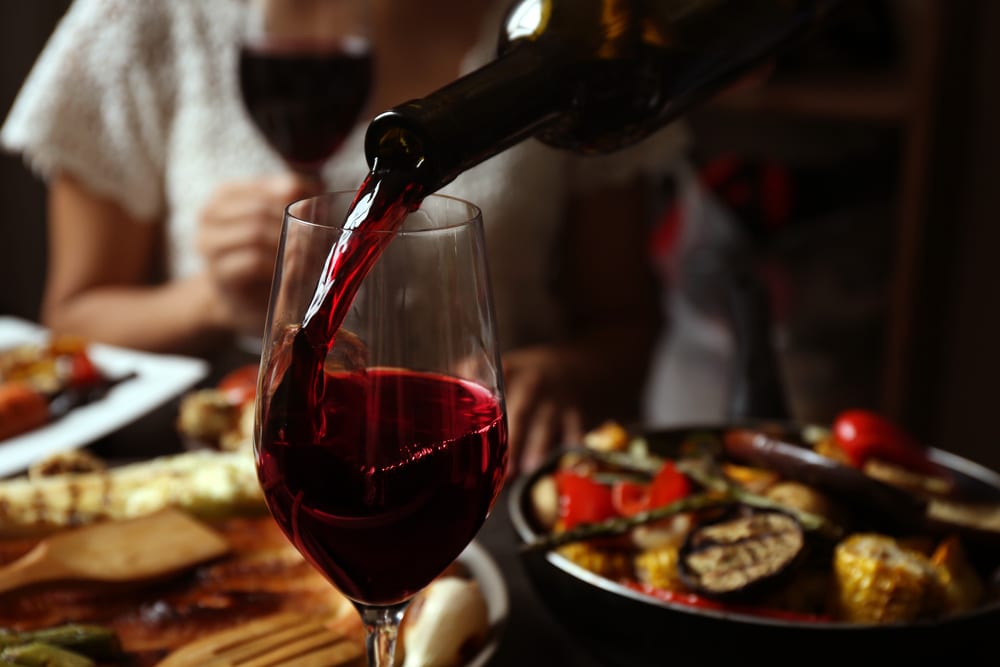 Glass of red wine being poured, with a bowl of roasted veg in the background